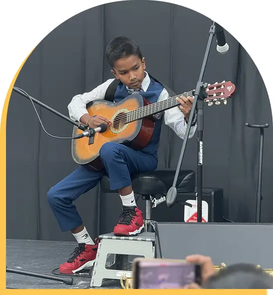 A young boy in a blue suit and red sneakers plays an acoustic guitar on stage, seated on a stool—a perfect showcase for music lessons Westchase families love. A microphone and music equipment are nearby as an audience member snaps a photo.