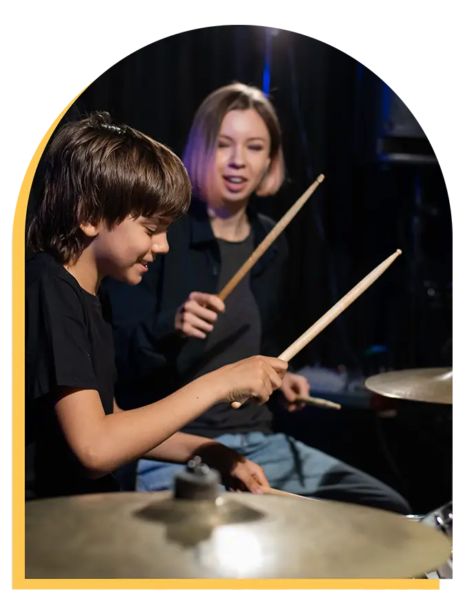 A smiling boy plays the drums while a woman behind him, also holding drumsticks, encourages him. Both appear to be enjoying music lessons in Westchase in a dimly lit room.