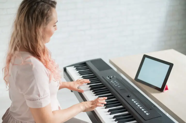 A woman with wavy hair plays an electronic keyboard while looking at a tablet on a stand. She is seated at a light-colored desk in a bright room with a white brick wall, capturing the inspiring environment often found during Music Lessons in East Orlando.