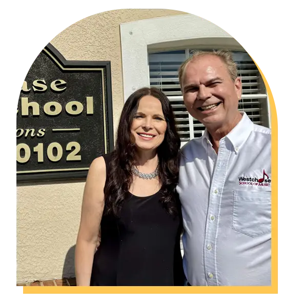 A smiling woman in a black dress and a smiling man in a white collared shirt stand together outside a building with a sign that includes "School" and the street number, hinting at music lessons Westchase might offer.