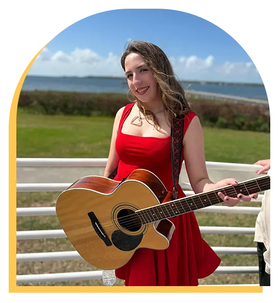 A smiling woman in a red dress stands outdoors by a white railing, holding an acoustic guitar—perfectly capturing the joy of music lessons Westchase has to offer. Behind her are green grass, water, and a blue sky dotted with clouds.